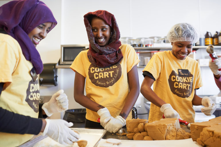 Careers & Technology in OST: Three smiling teens wearing "Cookie Cart" yellow t-shirts prepping cookie dough in a restaurant kitchen.