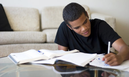 Teen wearing black t-shirt sits infront of white couch doing homework on coffee table.