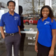VISTA: Two smiling young adults stand in front of glassdoor wearing blue polo shirts and slacks.