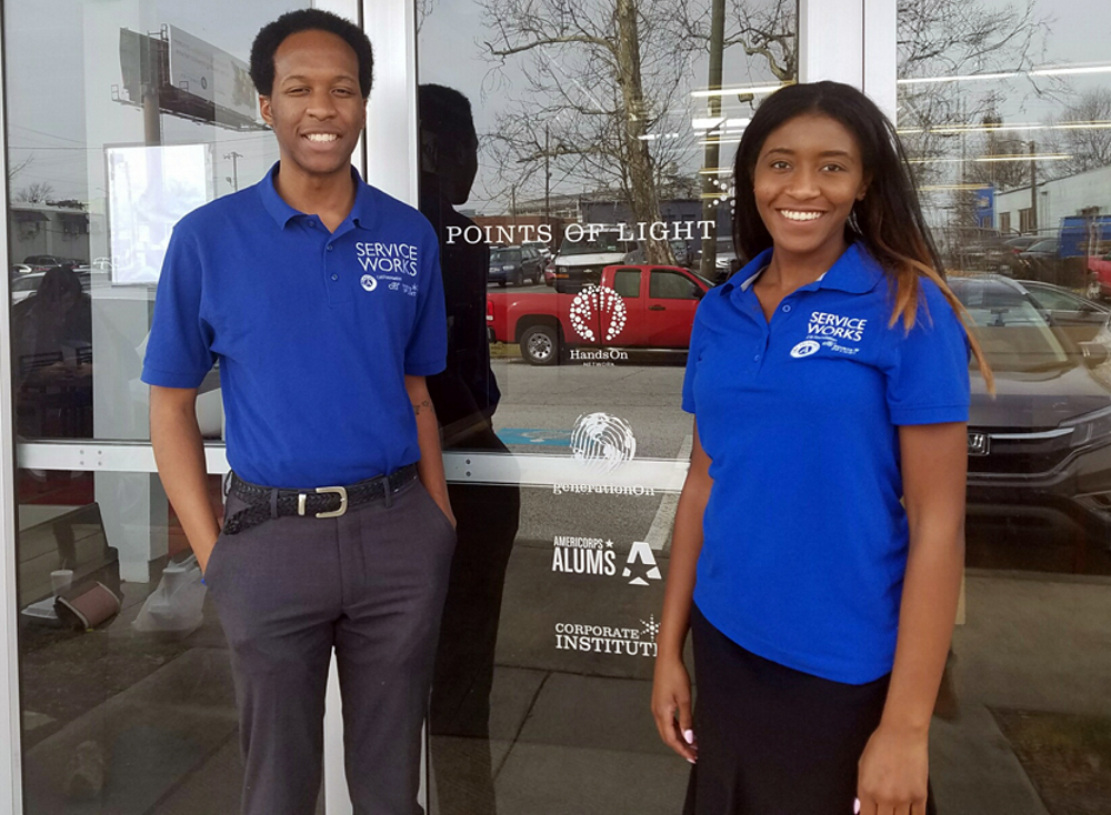 VISTA: Two smiling young adults stand in front of glassdoor wearing blue polo shirts and slacks.