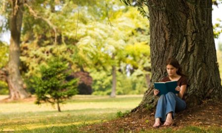 Learning anywhere, anytime: Middle school girls sits under huge green tree in park reading.