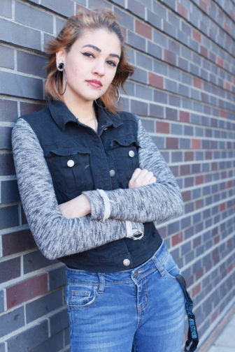Girl in jeans leaning on brick wall with arms crossed.