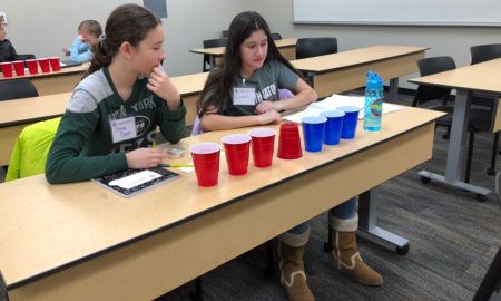 Two young girls with long brown hair in T-shirts, sitting at a long desk in a classroom, stare at empty blue and red cups in front of them, some turned up and some down.