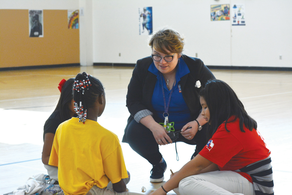 Afterschool activity room: Woman with short brown hair, dark, glasses wearing black blazer with rolled-up sleeves, blue shirt, dark pants and sneakers, squats down to talk to three girls in T-shirts sitting on the floor.