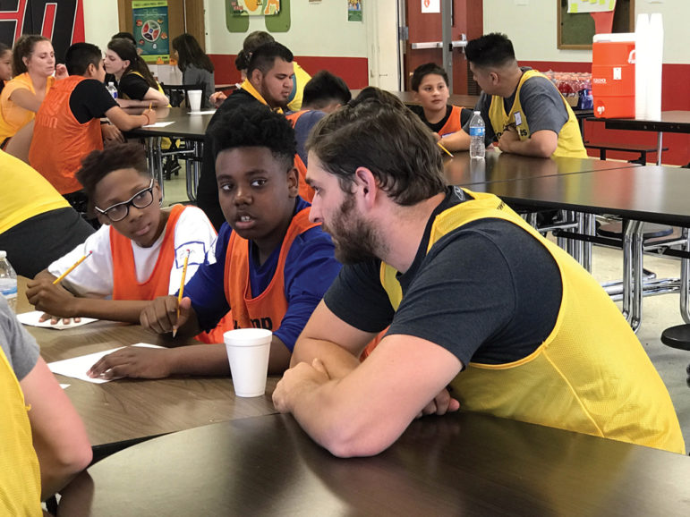 Afterschool program meeting in school cafeteria: Men wearing pinnies over T-shirts, looking serious, talk to boys and girls, also wearing pinnies over T-shirts, sitting at long tables together.