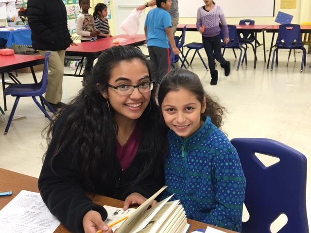 A young woman with long black hair and glasses smiles, working with a little girl with a brown ponytail as they sit at table with pencil and book.