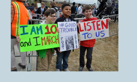 March for Our Lives: 3 boys hold up their signs; one in glasses and tan pants; one in dark jacket and jeans; one in red jacket and dark jeans.