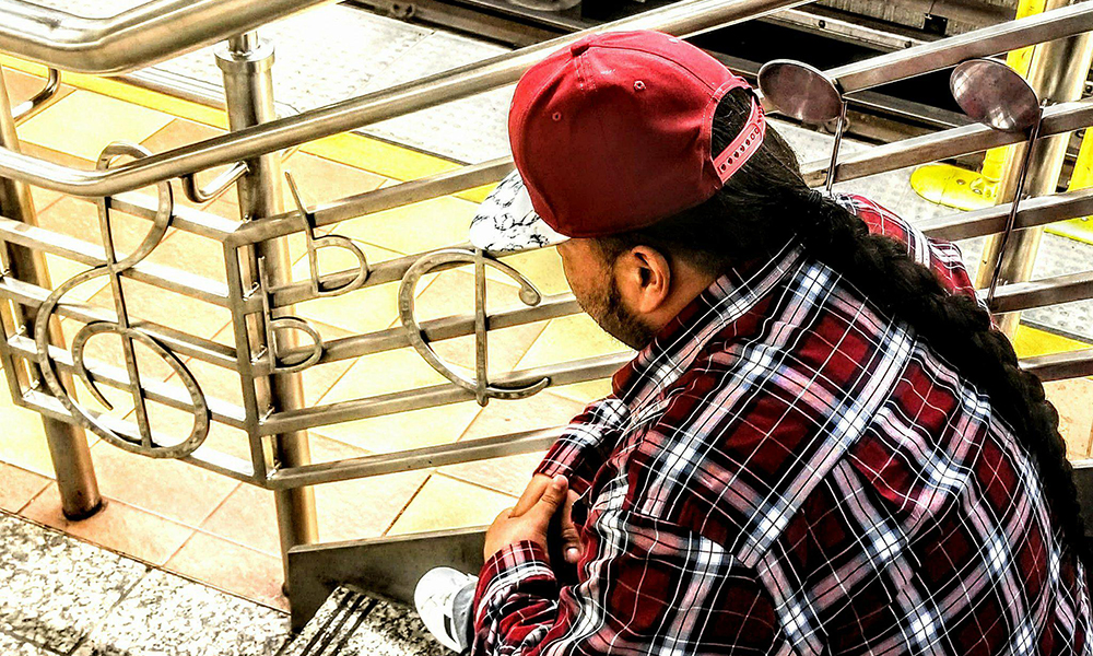 David in red ball cap, red plaid flannel shirt, long dark braid down his back, seen from back sitting on staircase with musical notes worked into balustrade.