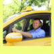 Smiling taxi driver in yellow cab, wearing beard, mustache, blue long-sleeved shirt with sleeves rolled up.