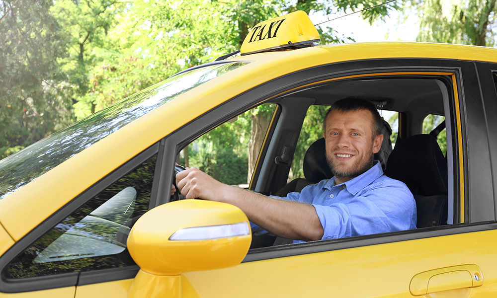 Smiling taxi driver in yellow cab, wearing beard, mustache, blue long-sleeved shirt with sleeves rolled up.