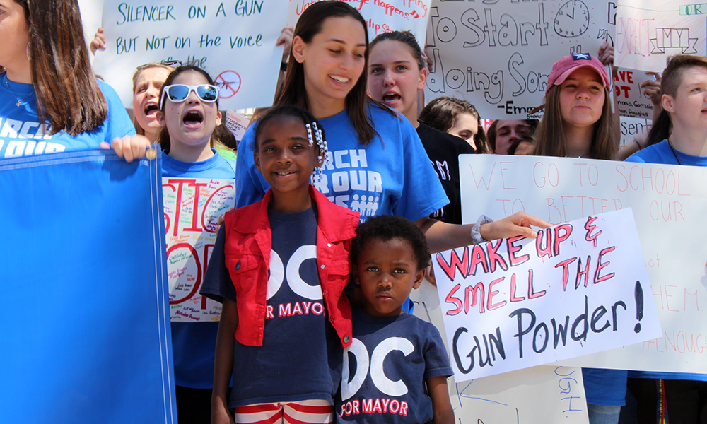  March for Our Lives: A little girl, wearing a DC for Mayor blue T-shirt, red and white striped pants and a blue vest, and a little boy wearing same T-shirt stand in front of older protestors at march.