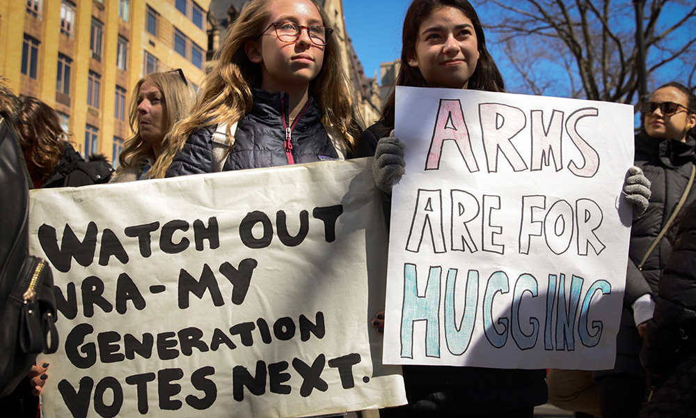 March for Our Lives: 2 young women hold up signs, one on left looks serious, wearing big glasses and black jacket; one on right smiles.