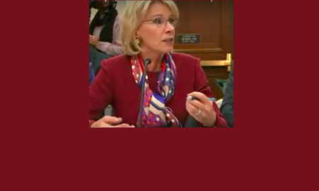 Education Secretary Betsy DeVos seated speaking at House Appropriations Committee wearing deep red suit & red/white/blue scarf.