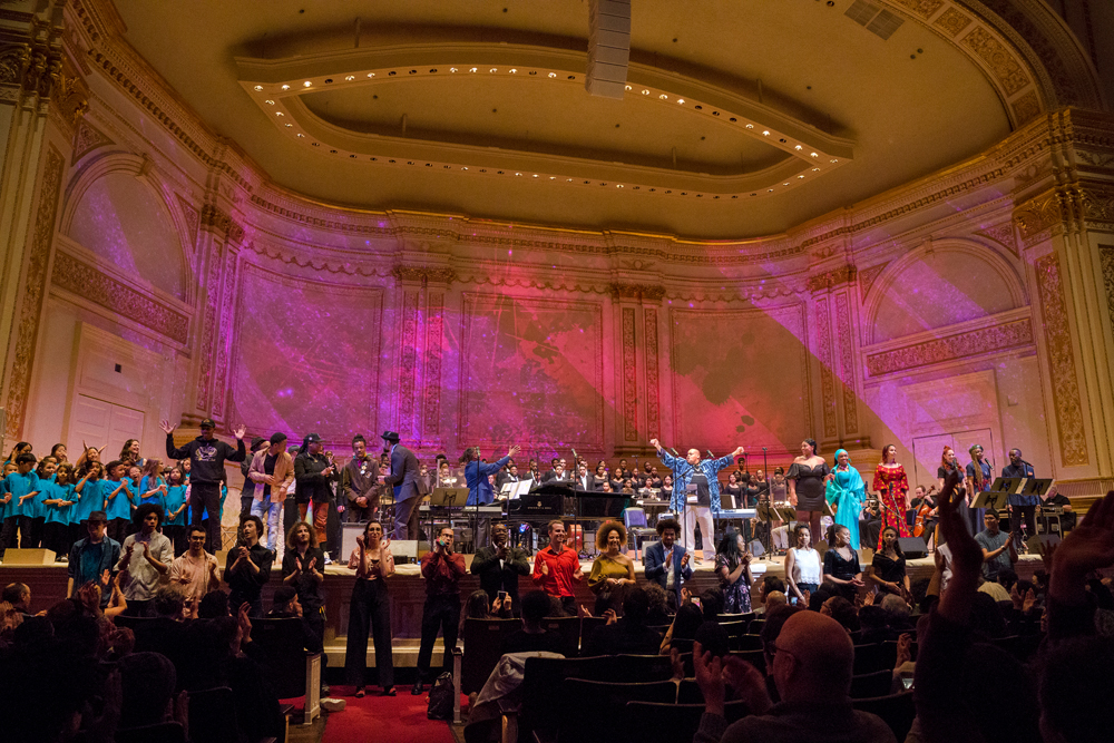 Orchestra, young people, young adults crowd stage in multicolored outfits, all performing at once.