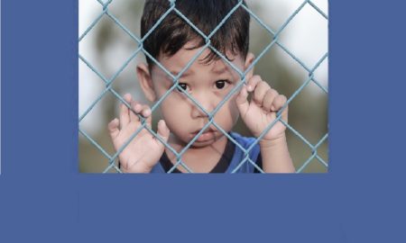 Foster care: Young boy sadly peering through chain link fence with hangs entwined in fence.