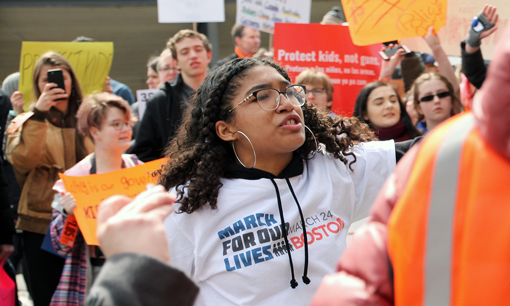 Student activist Laura-Luiza Gouvea in crowd of protesters, wearing March for Our Lives T-shirt, glasses and large circle earrings.