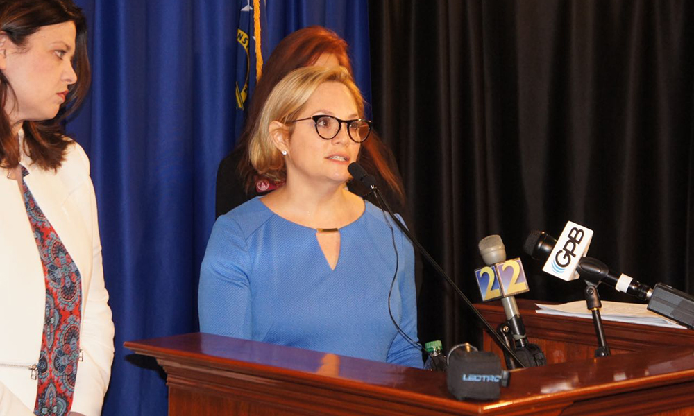  Child sexual abuse: Blonde woman in glasses, light blue dress speaks at podium with microphones from TV networks; side view of woman on her left with brown hair, white jacket, multicolored shirt.
