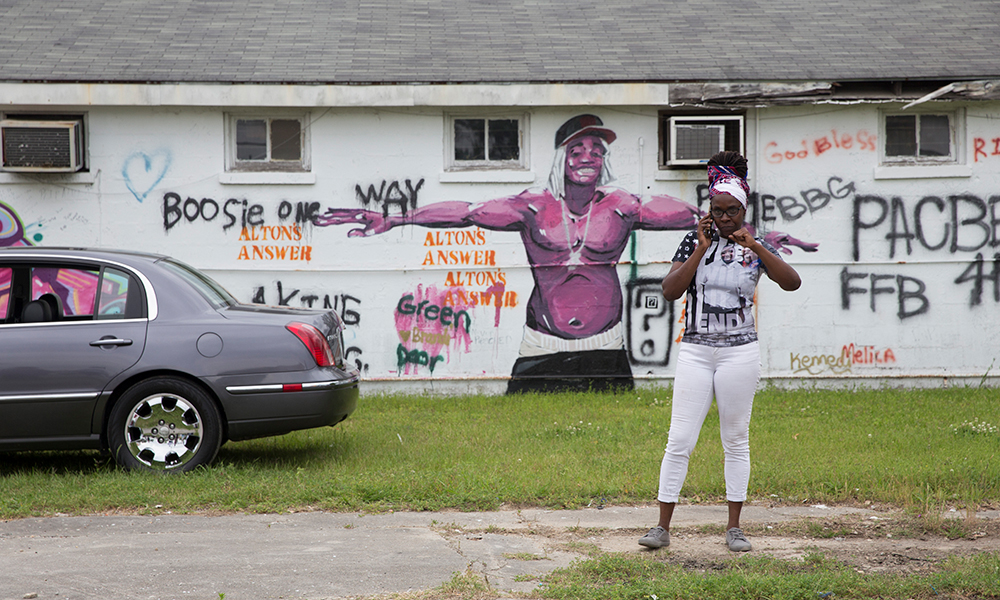 Mural of smiling shirtless man on side of building with cap, hanging ear coverings, underwear showing; graffiti surrounding him says Alton’s answer. Rear half of car on left side; woman with glasses, earrings, multicolored head wrap and multicolored top on phone on right.