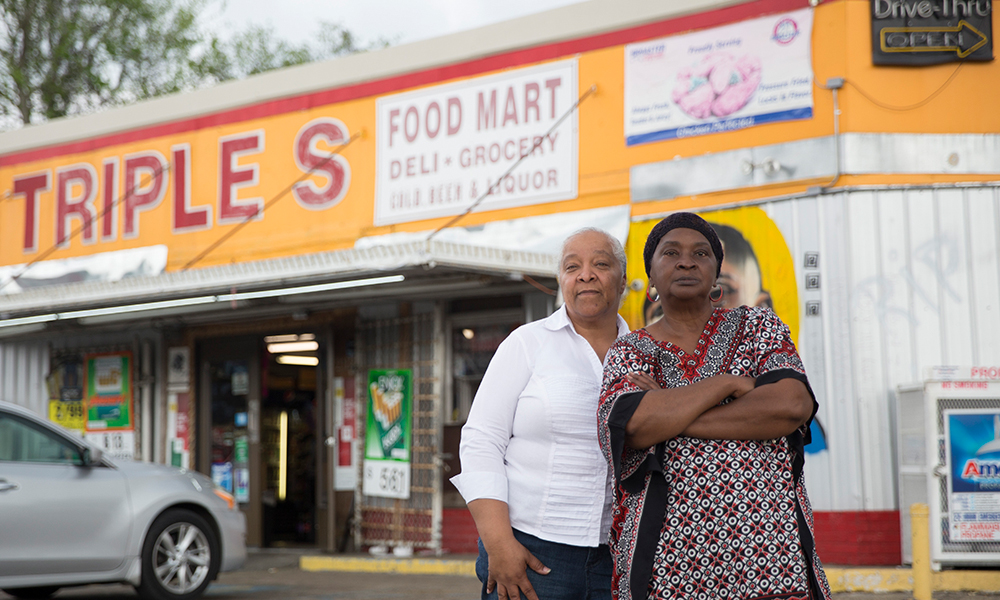 2 women, one in white shirt and dark pants, the other in a dashiki and black head wrap with her arms folded, stand in front of Triple S Food Mart.
