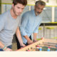 Adult play at work: young man in light beard, mustache, with green rubber band and green bracelet on right wrist and wearing gray T-shirt and jeans plays foosball with older man with heavier gray beard, mustache, wearing light blue shirt, dark pants