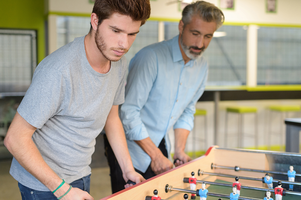 Adult play at work: young man in light beard, mustache, with green rubber band and green bracelet on right wrist and wearing gray T-shirt and jeans plays foosball with older man with heavier gray beard, mustache, wearing light blue shirt, dark pants