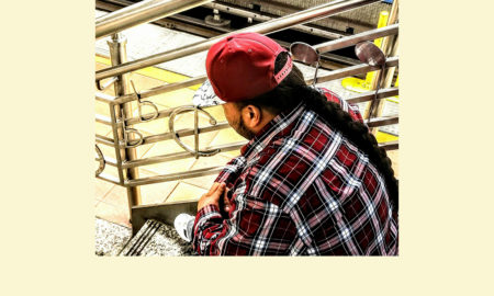 David in red ball cap, red plaid flannel shirt, long dark braid down his back, seen from back sitting on staircase with musical notes worked into balustrade.