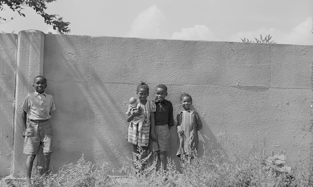 4 smiling children of color stand smiling against wall twice their height in 1941 black and white photo.