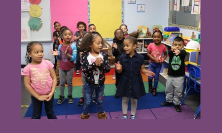 Locke Wellness Center class of children 4½ to 5 years old, most in T-shirts and pants, one in a dress and leggings, sing their welcome song in the morning standing on a multicolored carpet.