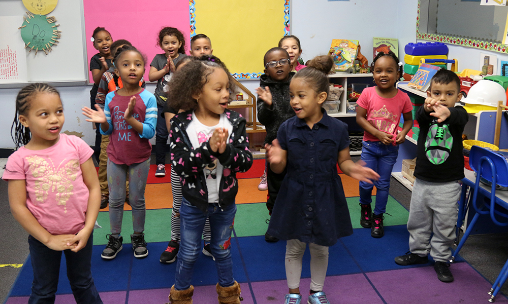 A class of children 4½ to 5 years old, most in T-shirts and pants, one in a dress and leggings, sing their welcome song in the morning standing on a multicolored carpet.