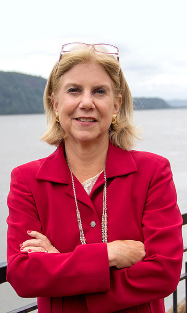 Shelley Mayer (headshot), blonde woman in red jacket, smiles against backdrop of river.