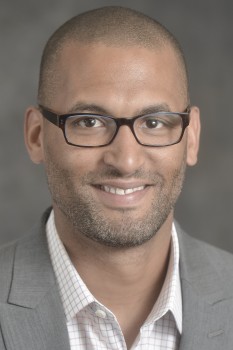 Michael Lens, an associate professor of urban planning and public policy at UCLA (headshot), smiling man with close buzz cut, 5 o’clock shadow beard and mustache wearing glasses, gray jacket, checked shirt