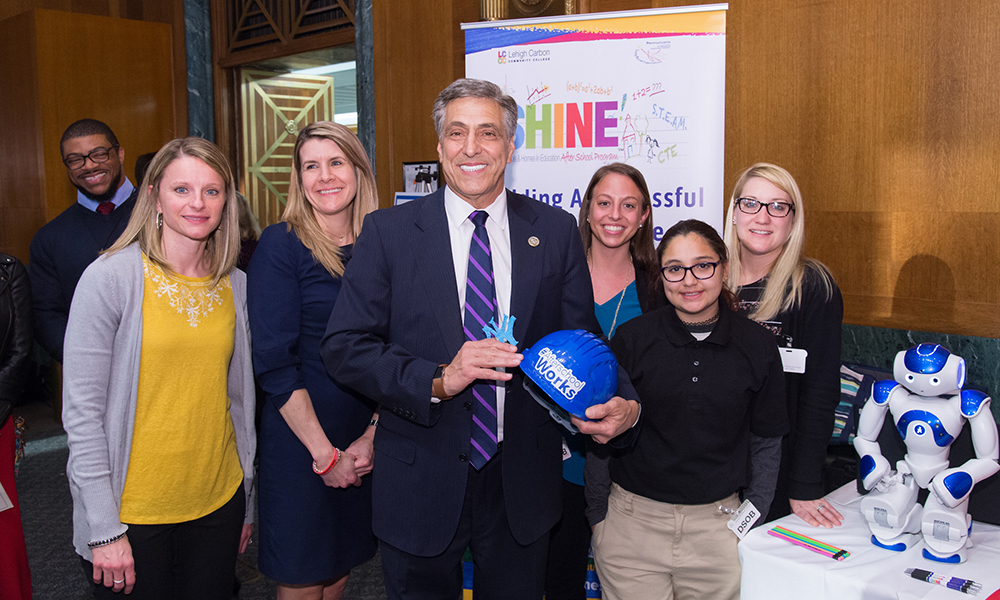 Afterschool Alliance: Tall smiling man in blue suit, striped tie, white shirt holding blue hard hat that says Afterschool Works surrounded by 5 smiling women.