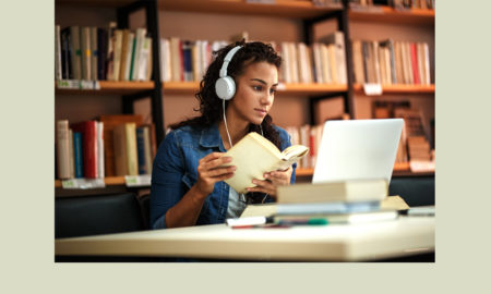 Personalized learning: Young woman in denim jacket with long dark hair wearing white headphones seated at table looks at white laptop. Bookshelves behind her.