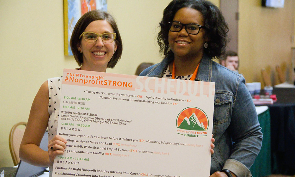 Two woman holding large poster board with agenda, wearing glasses and smiling at camera. 