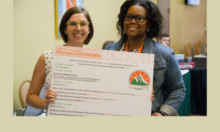 Two woman holding large poster board with agenda, wearing glasses and smiling at camera.