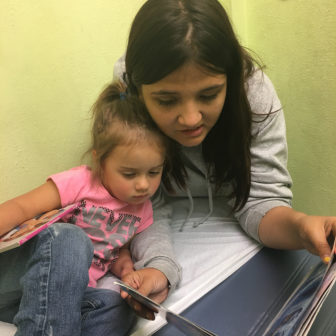 Foster care: Little girl in pink T-shirt, jeans, cuddles up to older girl in gray hoodie reading her a book.