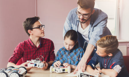Mentoring: 2 boys, 1 girl seated at table playing with vehicles with wires attached; man in glasses, open shirt over T-shirt bends over to point at paper on table.
