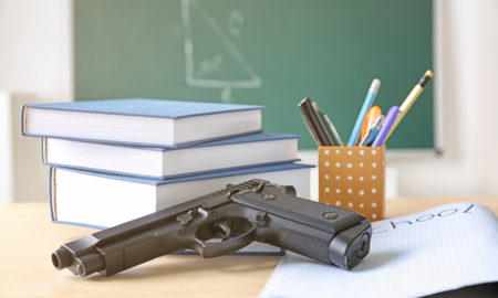 Gun violence: Gun lying on desk with 3 blue-covered books, paper with word School on it and pencil holder with pens, pencils, ruler; geometry drawing on blackboard is in background.
