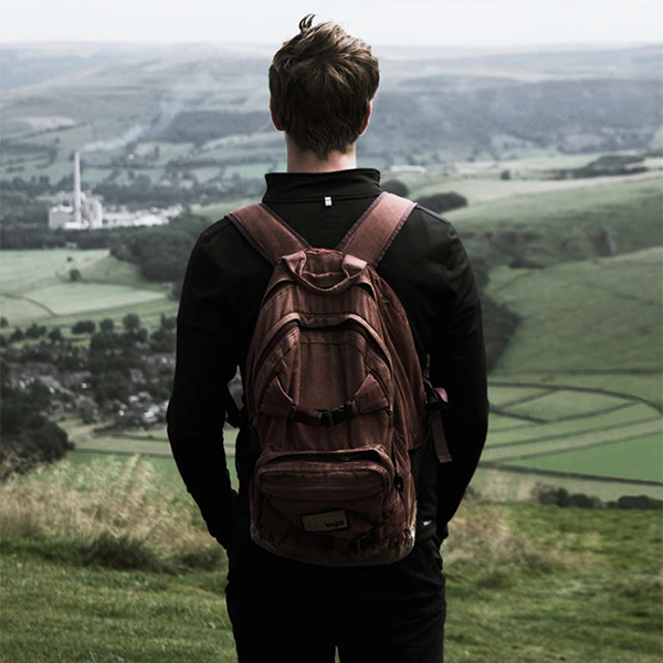 Homelessness: Young man wearing backpack stands at top of hill gazing down at green vista.