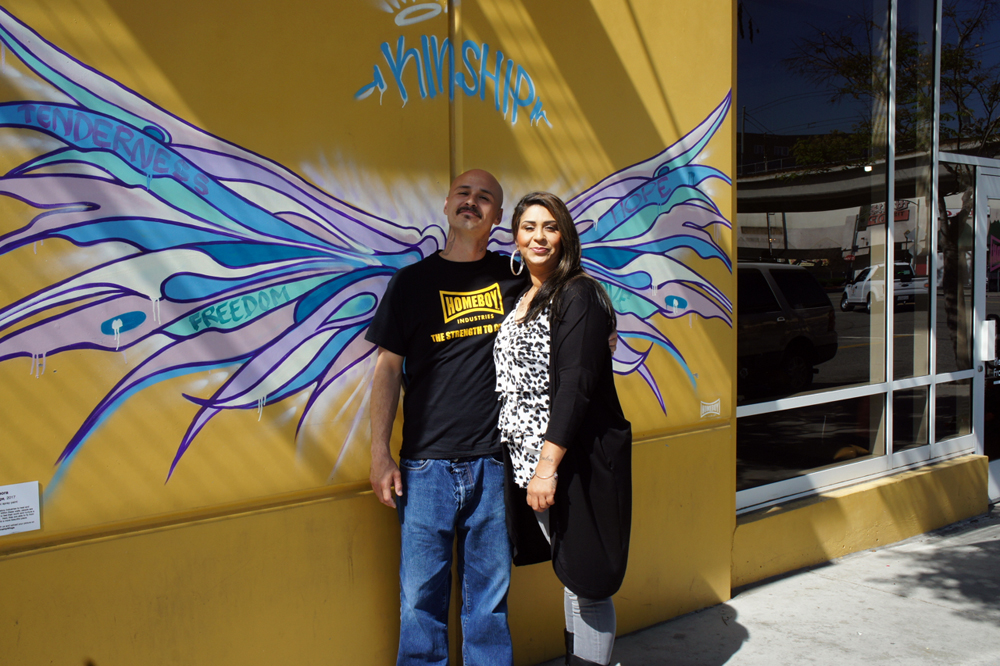 Gangs: Man and woman stand in front of yellow wall with mural of long angel wings. Man wears black T-shirt with Homeboy in yellow on it.
