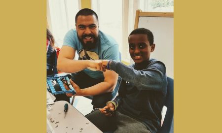 summer learning: Bearded man in T-shirt, boy in blue long-sleeved top tap fists as boy holds up item he’s been working on.