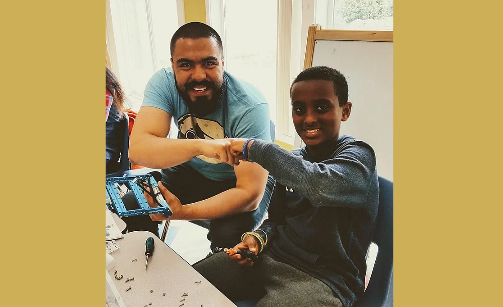 summer learning: Bearded man in T-shirt, boy in blue long-sleeved top tap fists as boy holds up item he’s been working on.