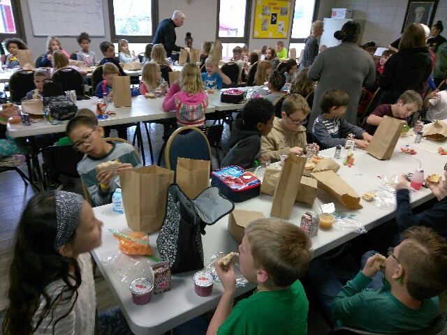 summer meals: children eat food from paper bags at long tables.