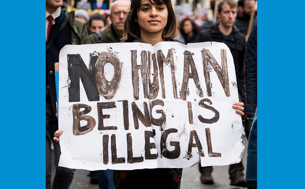Woman in crowd of protesters holds sign that says No human being is illegal.