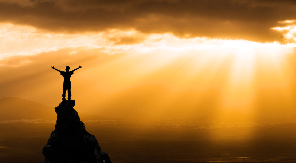 currency for youth success: person on top of rock with outstretched arms greeting sun - success concept
