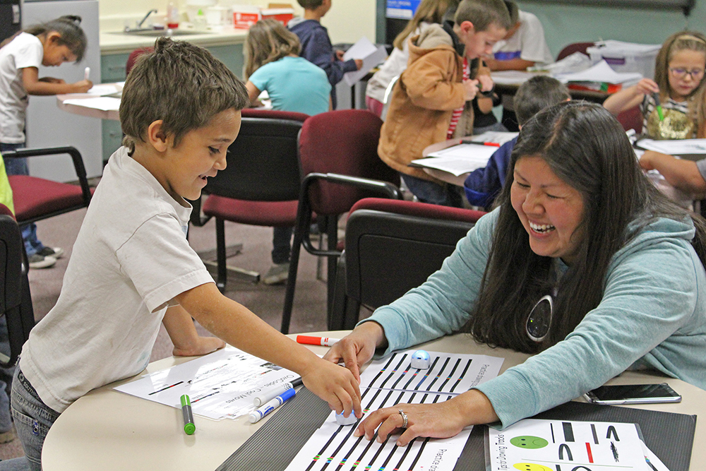 STEM: In classroom, smiling boy, woman lean over table and work together.