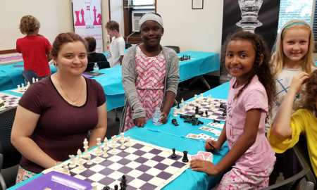 chess: Woman in brown shirt sits at chess board, surrounded by 3 smiling little girls.