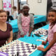 chess: Woman in brown shirt sits at chess board, surrounded by 3 smiling little girls.