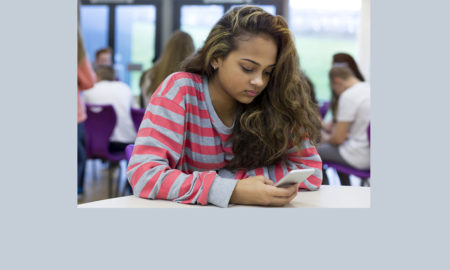 self-image: Female student sitting on her own at school. She has a smartphone in her hand and a stressed expression on her face.