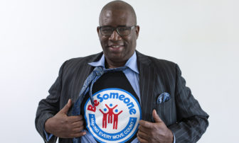 Orrin C. Hudson (headshot), founder and CEO of Be Someone, smiling man with short hair, glasses, holding open dark pinstriped jacket and light blue shirt to display T-shirt with Be Someone logo and motto on it